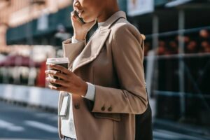 Mulher em traje formal caminha pela cidade enquanto conversa ao celular e segura uma xícara de café