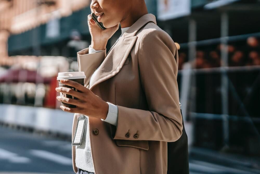 Mulher em traje formal caminha pela cidade enquanto conversa ao celular e segura uma xícara de café