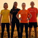 Quatro corredores exibem as camisetas oficiais da Maratona Internacional de Maringá em campo aberto ao pôr do sol