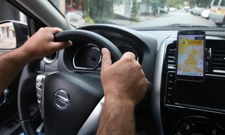 Motorista com as mãos ao volante dirige por rua arborizada, com celular apoiado no painel