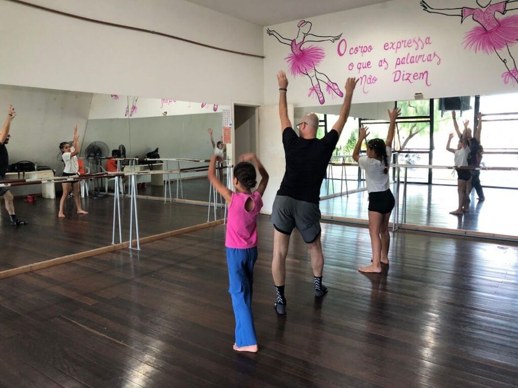 Um homem e crianças praticam dança em estúdio com espelhos e frases decorativas nas paredes