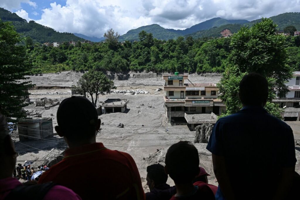 Pessoas observam área devastada por enchentes com edifícios parcialmente destruídos e terreno coberto de lama entre montanhas