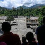Pessoas observam área devastada por enchentes com edifícios parcialmente destruídos e terreno coberto de lama entre montanhas
