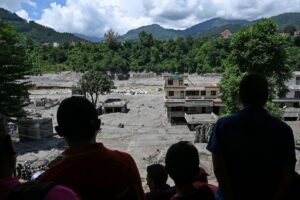 Pessoas observam área devastada por enchentes com edifícios parcialmente destruídos e terreno coberto de lama entre montanhas