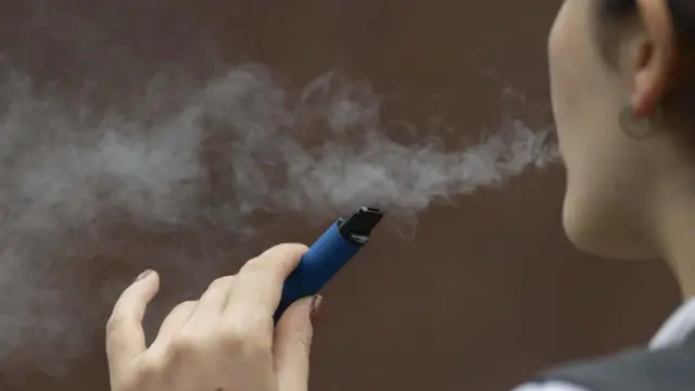 Uma pessoa segura um dispositivo eletrônico azul enquanto exala vapor em ambiente interno