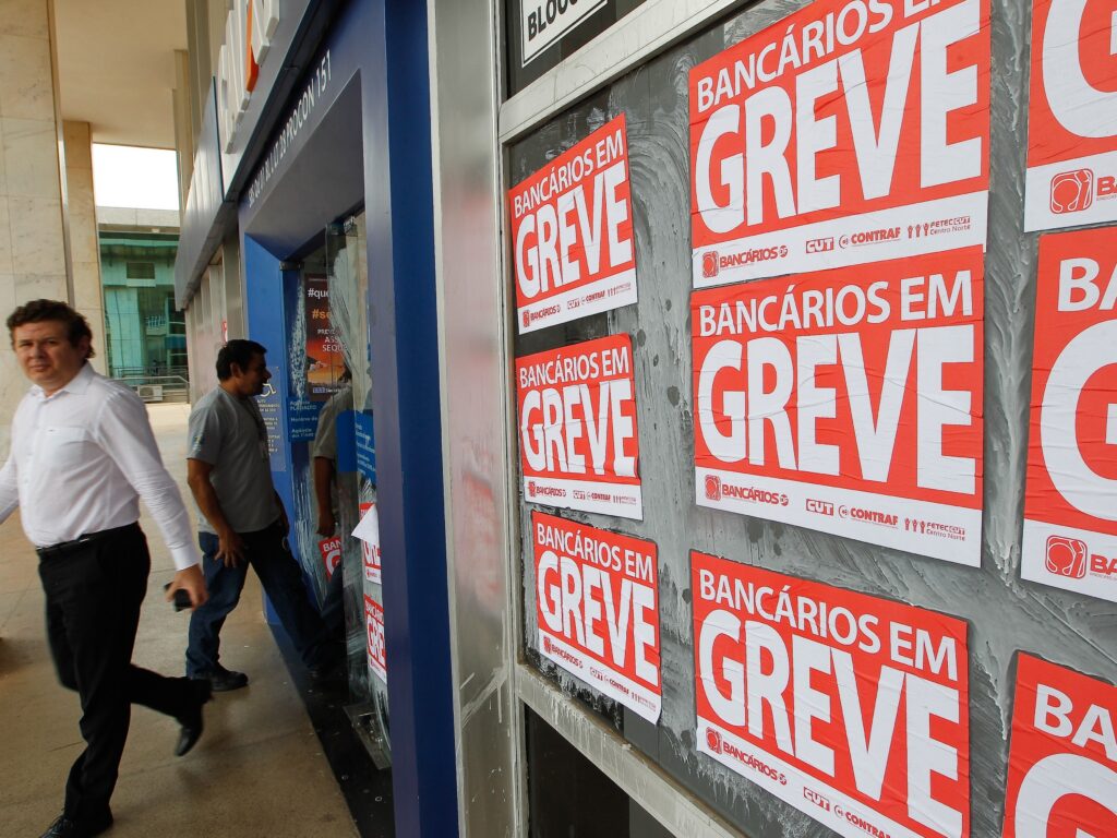 Cartazes de greve de bancários cobrem a fachada de uma agência, enquanto pessoas caminham pela calçada