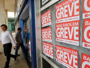 Cartazes de greve de bancários cobrem a fachada de uma agência, enquanto pessoas caminham pela calçada