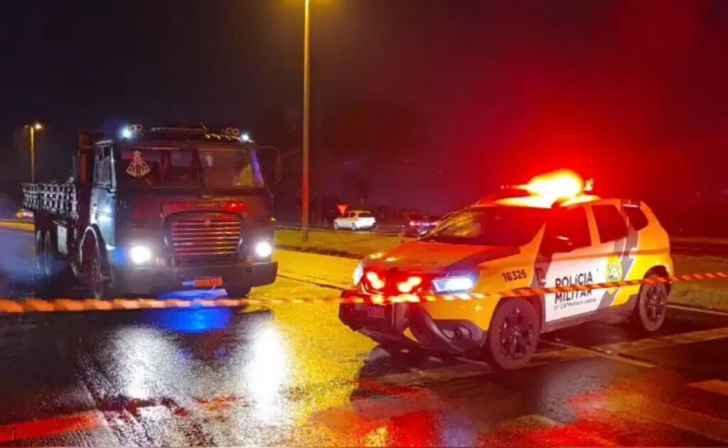 Caminhão danificado e veículo da Polícia Militar em rodovia à noite com faixa de isolamento
