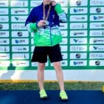 Jovem em pódio segura medalha com fundo de painel com logos de eventos esportivos