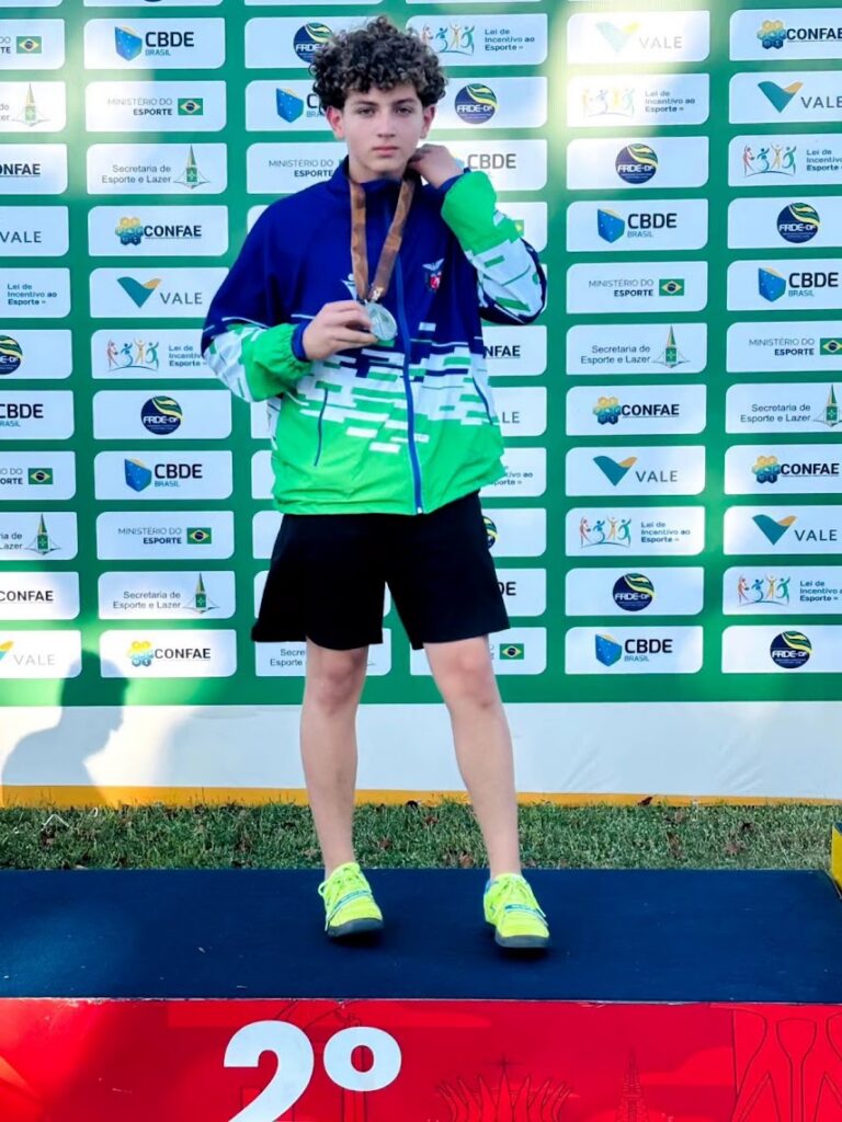Jovem em pódio segura medalha com fundo de painel com logos de eventos esportivos