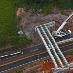 Vista aérea de obras em rodovia com estrutura de ponte sendo montada e veículos circulando ao redor