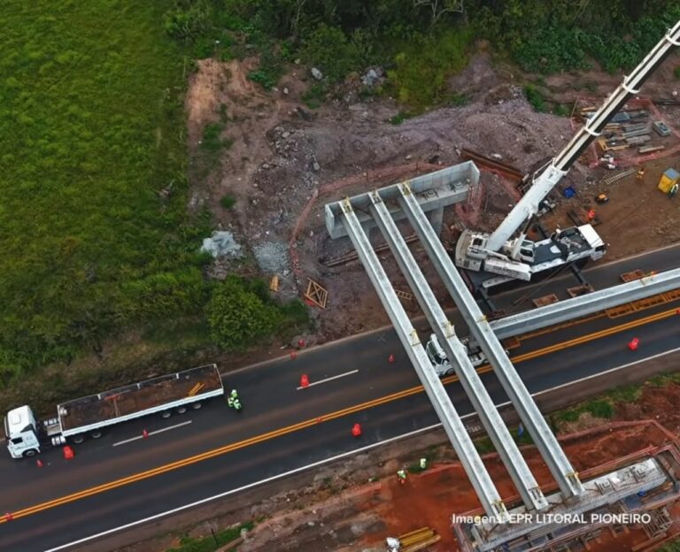 Vista aérea de obras em rodovia com estrutura de ponte sendo montada e veículos circulando ao redor