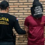 Um homem com capuz é conduzido por agente da Polícia contra o Crime Organizado em frente a parede de tijolos