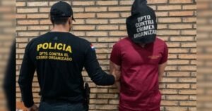 Um homem com capuz é conduzido por agente da Polícia contra o Crime Organizado em frente a parede de tijolos