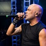 Um homem calvo canta no palco com microfone em frente a público