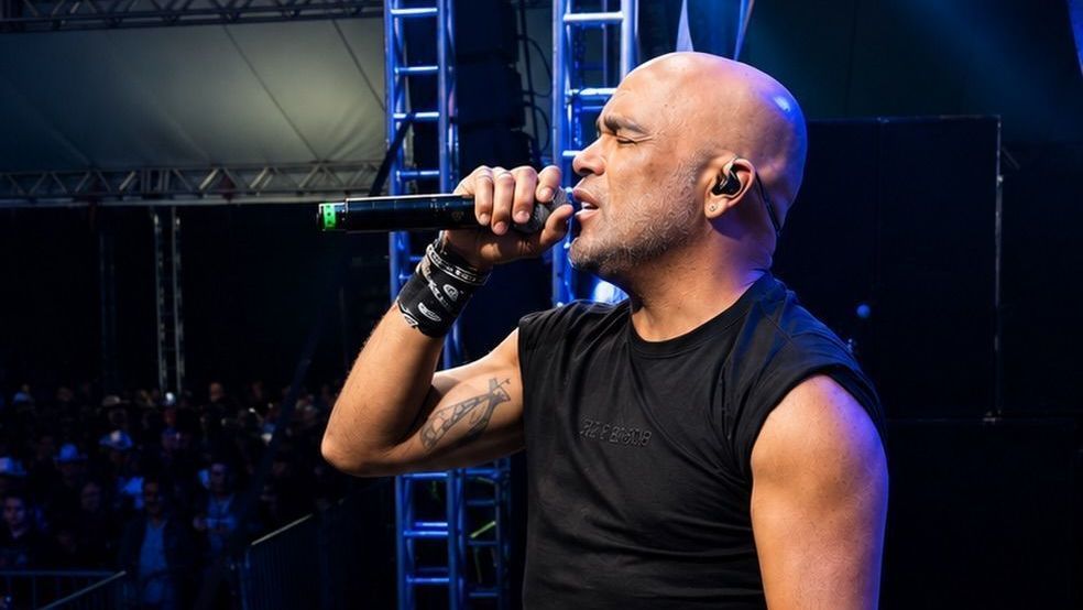 Um homem calvo canta no palco com microfone em frente a público