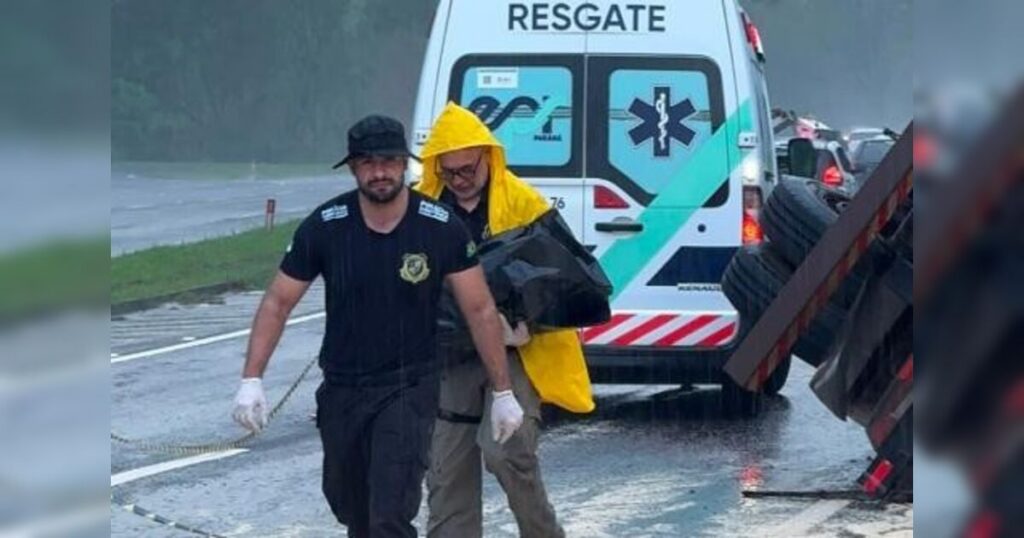 Dois homens carregam um corpo em saco preto sob chuva forte ao lado de veículo de resgate em rodovia molhada