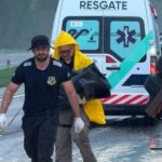 Dois homens carregam um corpo em saco preto sob chuva forte ao lado de veículo de resgate em rodovia molhada