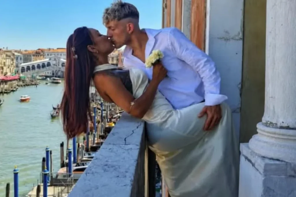 Um homem e uma mulher se beijam em um balcão com vista para um canal e uma ponte arqueada ao fundo