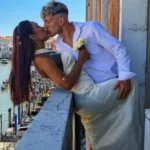 Um homem e uma mulher se beijam em um balcão com vista para um canal e uma ponte arqueada ao fundo