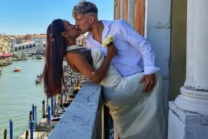 Um homem e uma mulher se beijam em um balcão com vista para um canal e uma ponte arqueada ao fundo