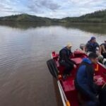 Quatro homens em barco vermelho navegam por represa cercada por colinas arborizadas