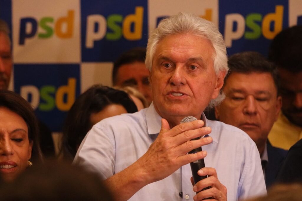Um homem de cabelos brancos fala com microfone em frente a painel com logotipo do PSD, cercado por pessoas