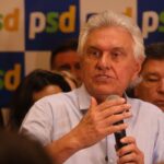 Um homem de cabelos brancos fala com microfone em frente a painel com logotipo do PSD, cercado por pessoas