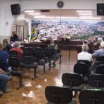Pessoas sentadas em cadeiras de uma sala com mesa elevada e bandeiras ao fundo