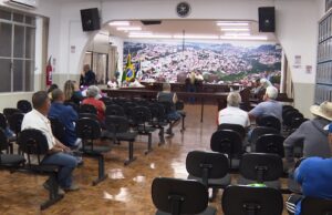 Pessoas sentadas em cadeiras de uma sala com mesa elevada e bandeiras ao fundo
