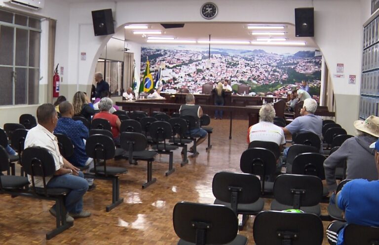 Pessoas sentadas em cadeiras de uma sala com mesa elevada e bandeiras ao fundo