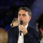 Um homem de jaqueta escura e camisa branca fala em microfone diante de público em evento noturno