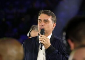 Um homem de jaqueta escura e camisa branca fala em microfone diante de público em evento noturno