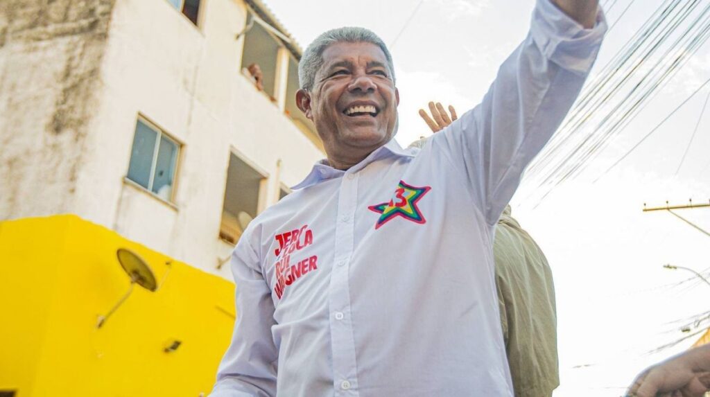 Um homem de camisa branca com estampa de estrela e nome sorrindo e acenando em frente a um prédio com parede amarela