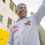 Um homem de camisa branca com estampa de estrela e nome sorrindo e acenando em frente a um prédio com parede amarela
