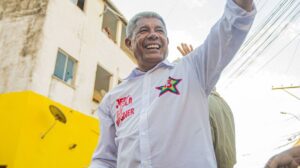 Um homem de camisa branca com estampa de estrela e nome sorrindo e acenando em frente a um prédio com parede amarela