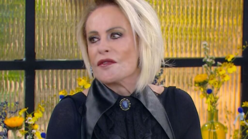 Uma mulher de cabelos claros e blusa escura em frente a um fundo com flores amarelas e painel dourado