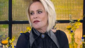 Uma mulher de cabelos claros e blusa escura em frente a um fundo com flores amarelas e painel dourado