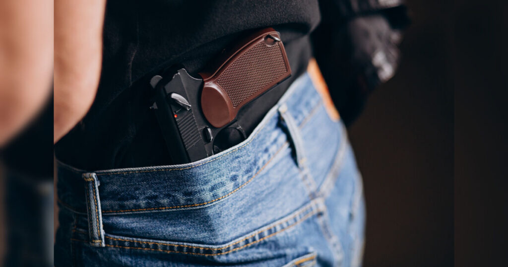 Pessoa com pistola presa na cintura, parcialmente visível sob camiseta e calça jeans