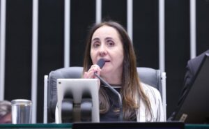 Uma mulher fala em microfone durante reunião em ambiente formal com painel de listras verticais ao fundo