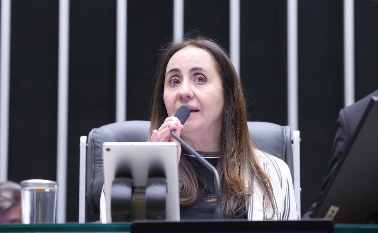 Uma mulher fala em microfone durante reunião em ambiente formal com painel de listras verticais ao fundo