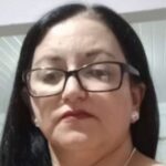 Mulher de cabelos escuros e óculos olha para a câmera em ambiente interno