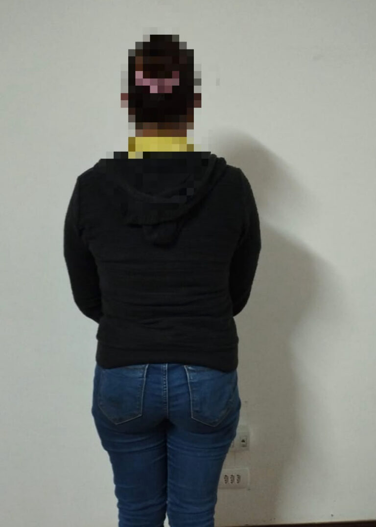 Uma mulher de costas, com moletom preto e calça jeans, em frente a parede branca