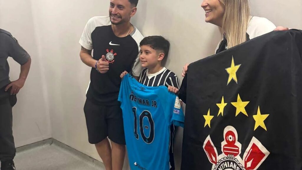 Um garoto segura camisa azul com o nome "Neymar Jr" ao lado de um homem e uma mulher, que exibem bandeira do Corinthians em ambiente interno
