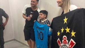 Um garoto segura camisa azul com o nome "Neymar Jr" ao lado de um homem e uma mulher, que exibem bandeira do Corinthians em ambiente interno