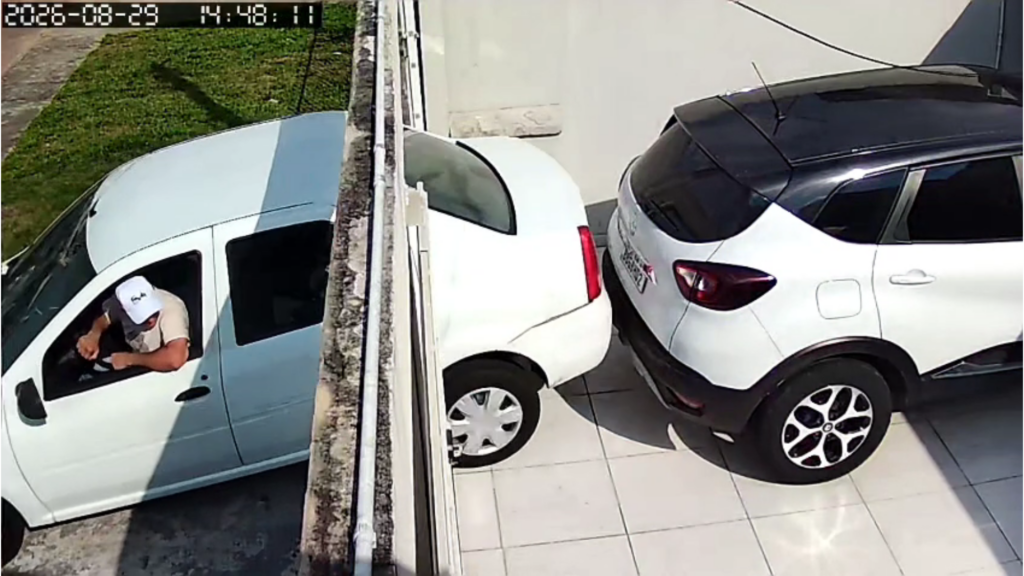 Um homem dentro de uma van branca com o braço para fora, próximo a dois carros estacionados em área externa
