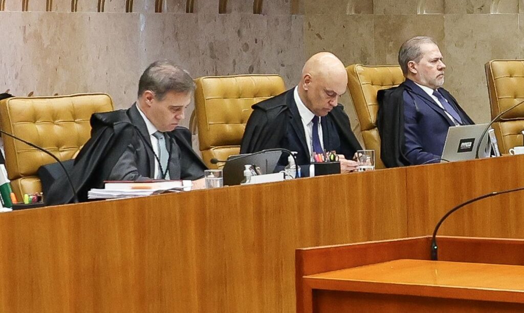 Três homens em trajes formais sentados em cadeiras douradas em um plenário com mesa de madeira e microfones