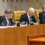 Três homens em trajes formais sentados em cadeiras douradas em um plenário com mesa de madeira e microfones