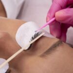 Mãos com luvas aplicam produto em cílios de pessoa deitada com protetores nos olhos