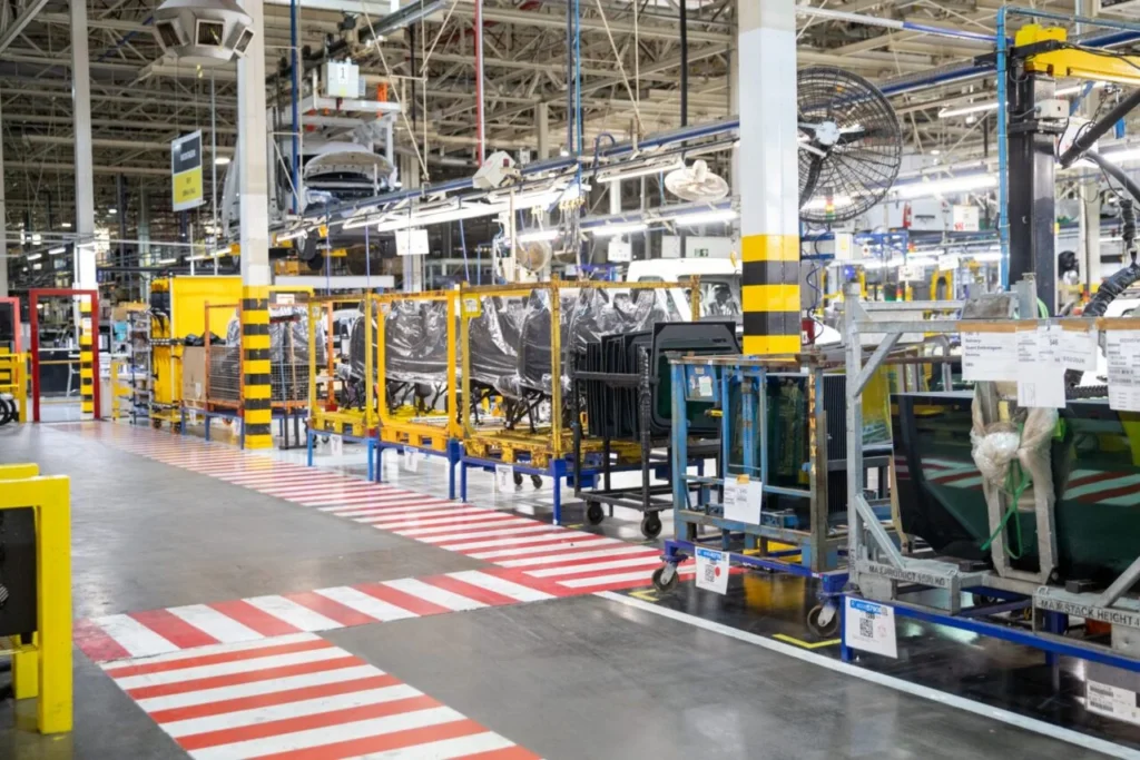 Interior de uma fábrica com esteiras de montagem, peças automotivas em carrinhos e estruturas metálicas suspensas